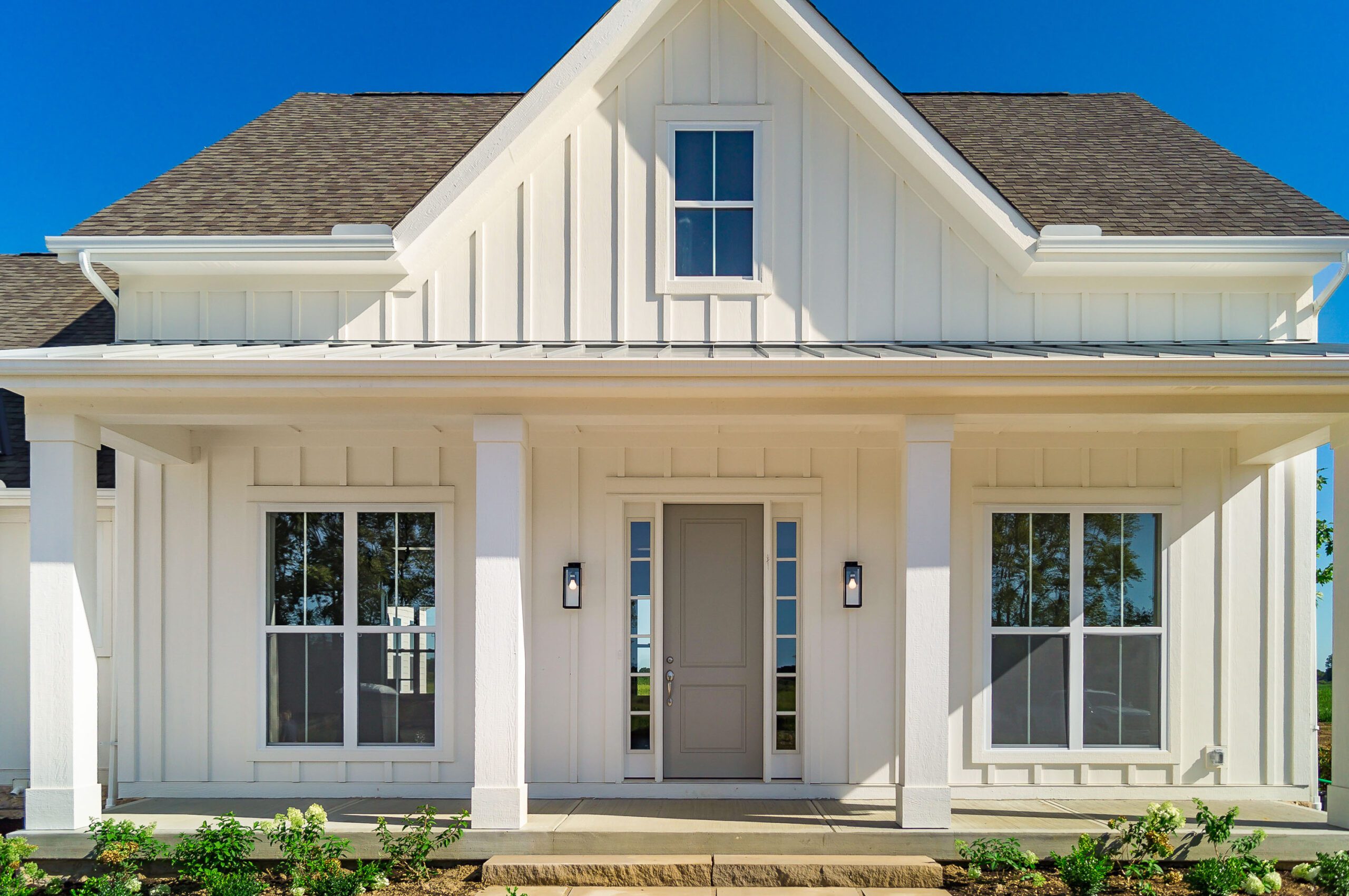 A white two-story house with a front entrance featuring a grey door, flanked by large windows and two exterior wall lights, with a porch supported by four columns.